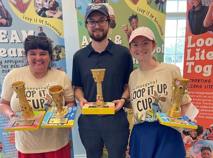 Nathan Durham stands with Loop It Up Members Molly and Erika. They are all holding trophies that were created by the kids supported by Loop It Up for their golf tournament